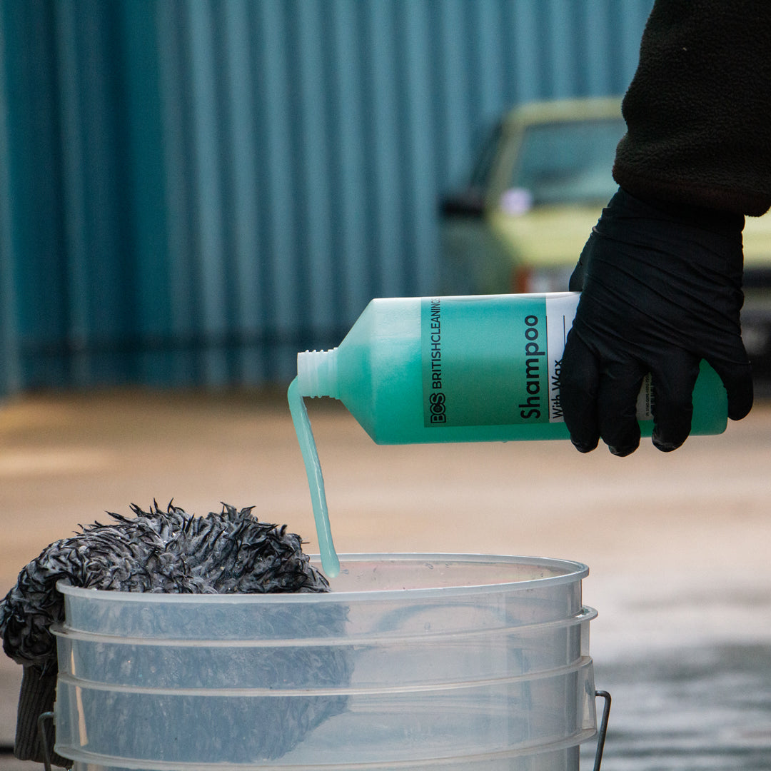 Person pouring shampoo from a bottle into a bucket with a car in the background